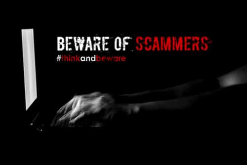A text "Beware of scammers. #think and beware" with low key motion blur male hand typing on keyboard laptop